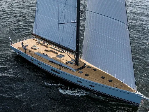 A sleek sailing yacht with large sails glides over calm waters featuring a spacious wooden deck and two individuals on board A sleek sailing yacht with large sails glides over calm waters, featuring a spacious wooden deck and two individuals on board.